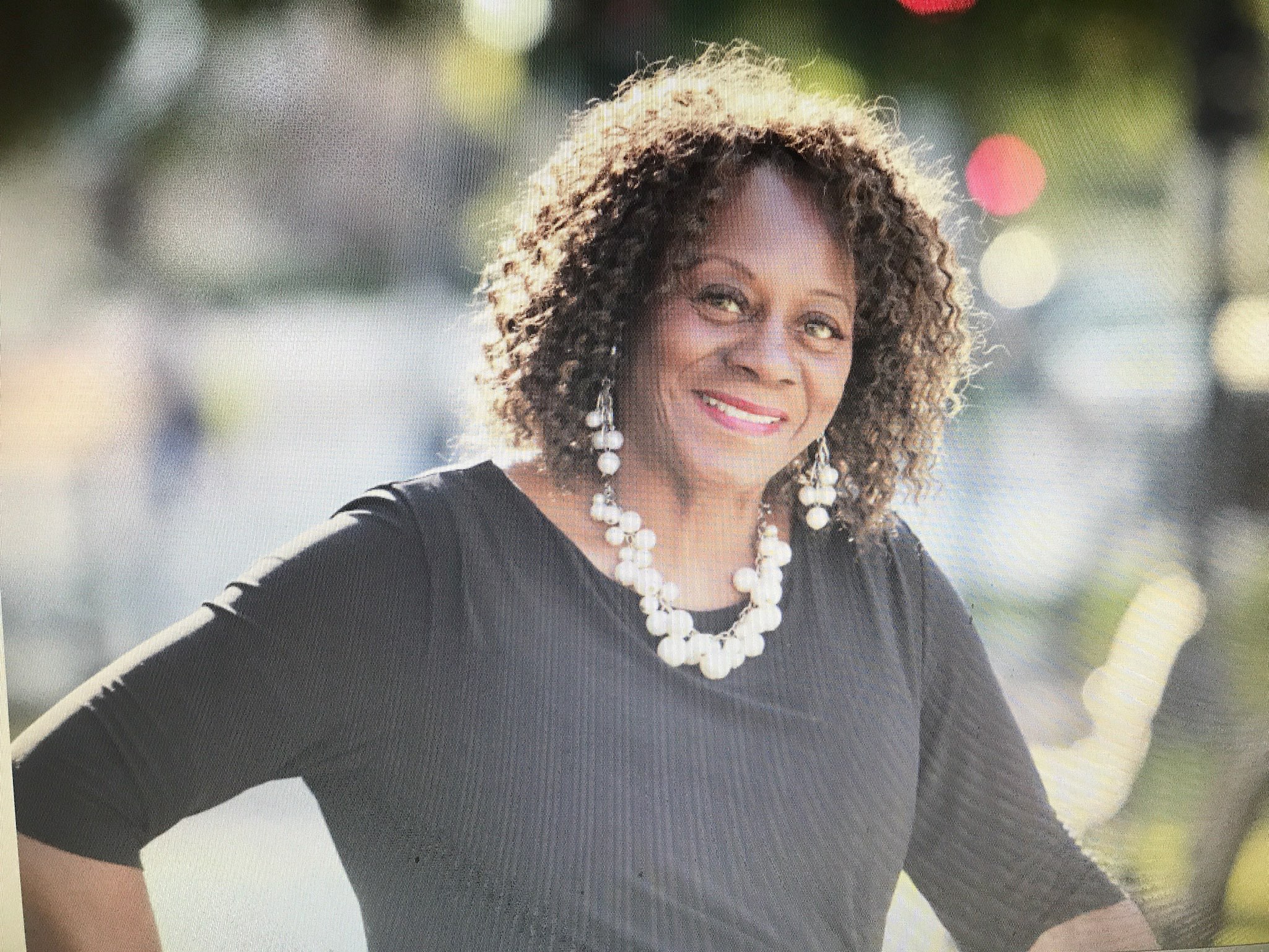 Portrait of Dee smiling outdoors in a black top with statement jewelry, in soft natural light.